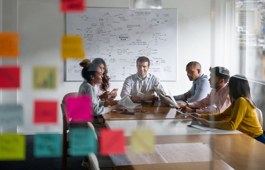 Six diverse professionals in a modern office meeting with sticky notes and a whiteboard in the background.
