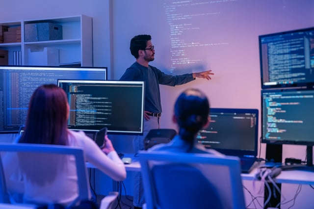 Instructor presenting code on a projected screen to a group of students in a computer lab, with multiple monitors displaying programming interfaces in a collaborative learning environment.