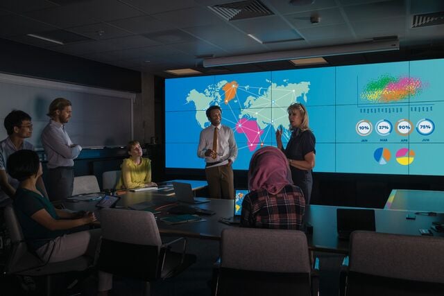 People in a conference room view a large screen displaying global data visualizations, including maps, charts, and graphs, during a collaborative presentation